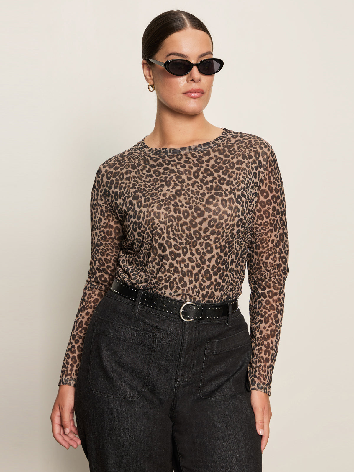 A woman wearing the Sanctuary Clothing long sleeve perfect mesh tee in French Kitty (extended sizing), black sunglasses, black belted pants, and hoop earrings stands confidently against a plain light background.