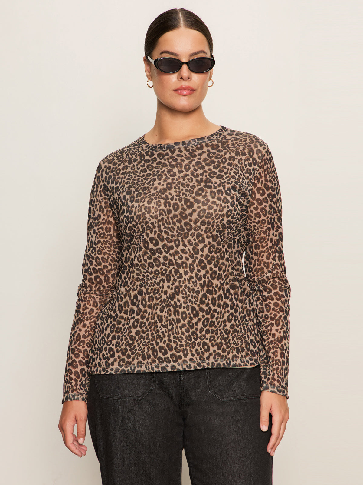 A woman with dark hair pulled back wears black sunglasses, gold hoop earrings, and Sanctuary Clothing's long sleeve perfect mesh tee french kitty (extended sizing), paired with dark pants, standing against a plain light background.