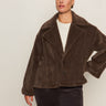 A woman wears the Sanctuary Clothing colette fur jacket choco chip extended sizing, featuring dark brown faux fur with large lapels and two buttons, over a white top and dark pants, standing against a plain light background.