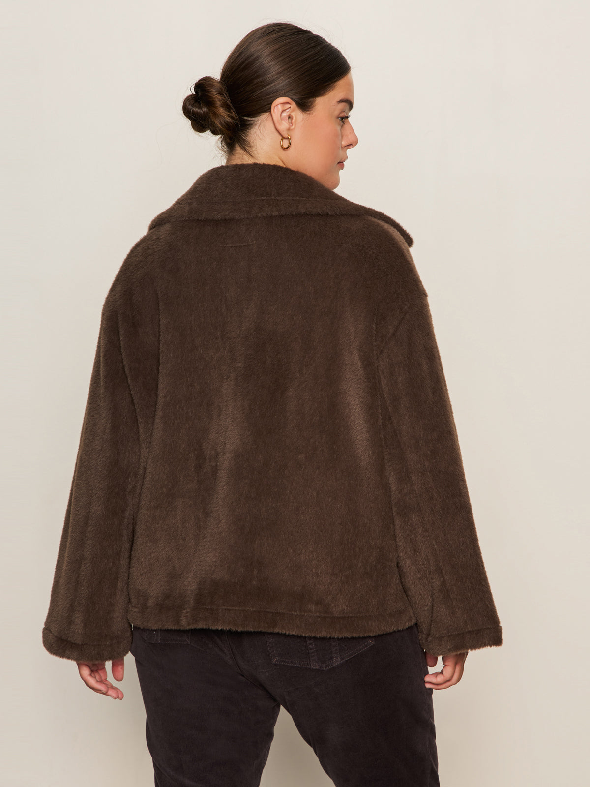 A woman with dark hair in a bun models the Sanctuary Clothing colette fur jacket choco chip (extended sizing) with black pants, facing away from the camera against a plain light background.