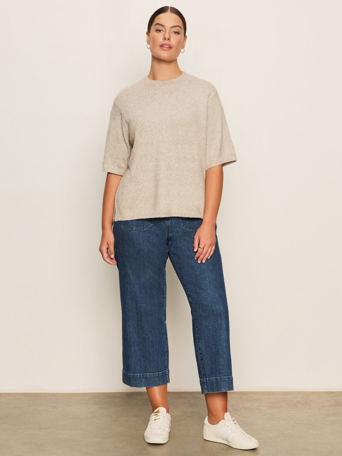 A woman stands against a plain light wall, wearing Sanctuary Clothing’s perfect sweater tee in heather pearl (extended sizing), blue wide-leg cropped jeans, and white sneakers. Her hair is pulled back and she looks at the camera with a relaxed posture.
