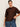 A woman with dark hair in a low bun wears gold hoop earrings, blue jeans, and the Sanctuary Clothing perfect sweater tee chicory extended sizing, standing against a light background and holding her sweater with one hand.