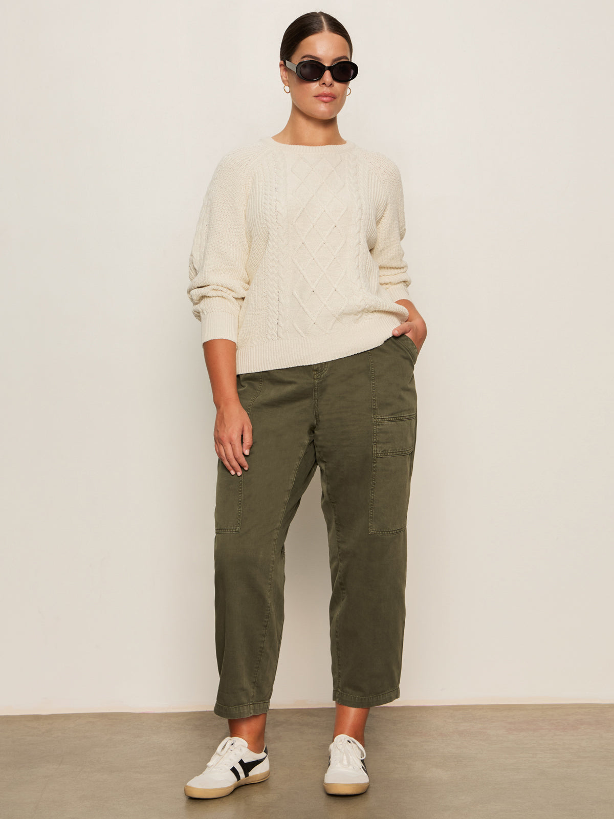 A person wearing the Sanctuary Clothing nomad cable popover chalk extended sizing sweater, olive green cargo pants, white sneakers, and black sunglasses stands against a plain white wall with one hand in their pocket.