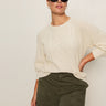 A woman wears Sanctuary Clothing's nomad cable popover in chalk (extended sizing) and black sunglasses, paired with olive green pants, posing against a light background with one hand on her hip.