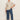 A woman poses against a plain background in Sanctuary Clothing’s the marine jeans aspen extended sizing—wide-leg blue jeans—paired with a beige short-sleeve sweater and white sneakers, her left hand in her pocket and a relaxed expression.