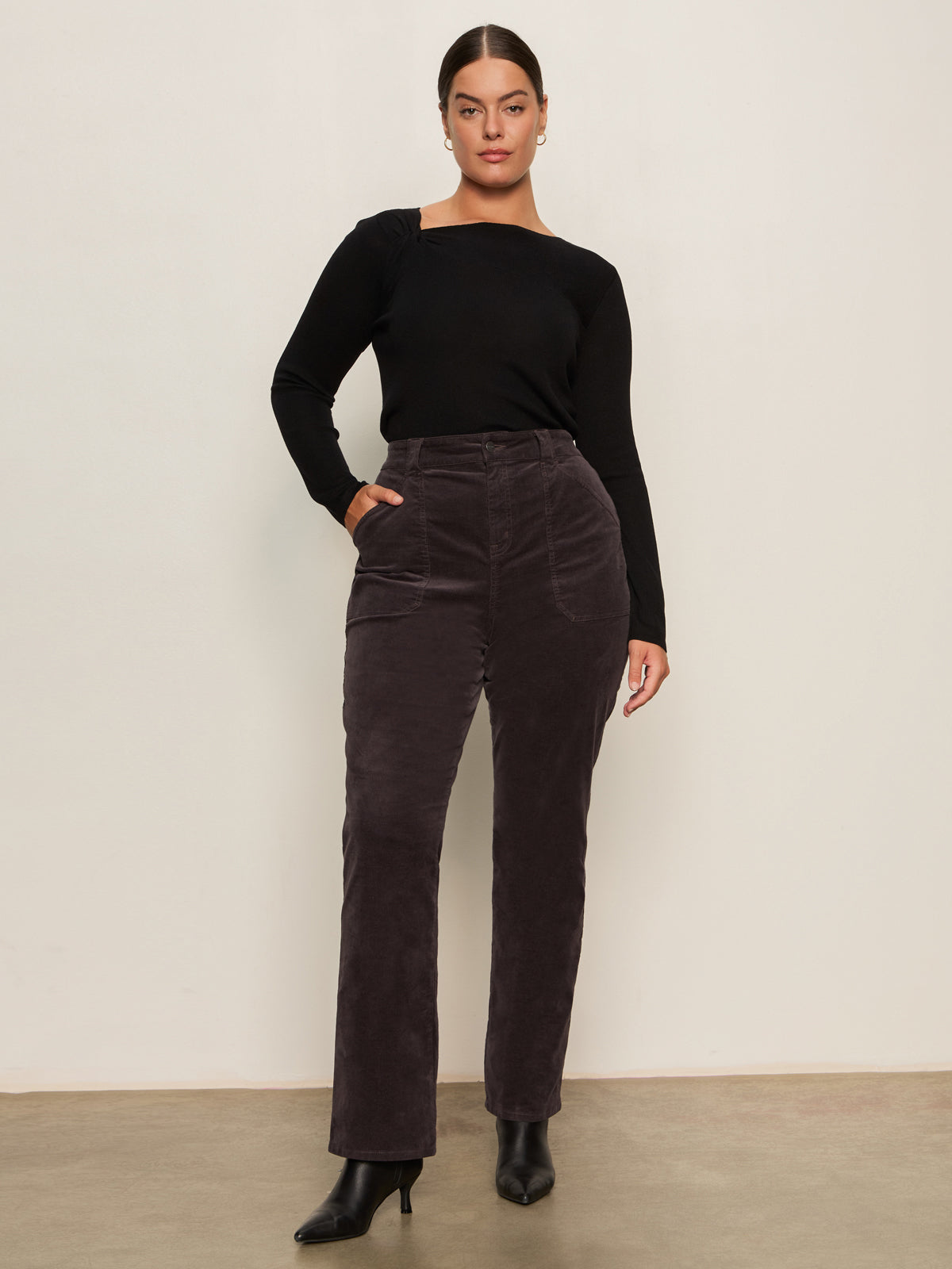 A woman with her hair pulled back and hand in pocket wears Sanctuary Clothing's corduroy hayden bootcut pant semi sweet extended sizing, paired with a fitted black top and black heeled ankle boots, standing against a light wall.