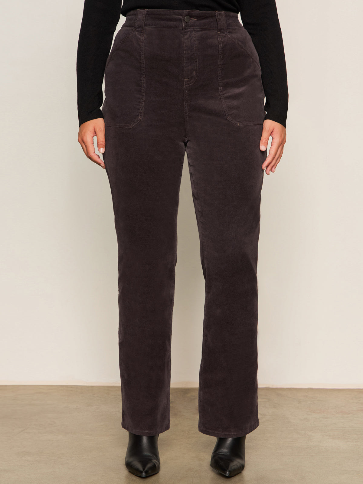 A person wears Sanctuary Clothing's Corduroy Hayden Bootcut Pant in Semi Sweet (extended sizing) with front patch pockets, paired with a black long-sleeve top and black ankle boots, against a light background.