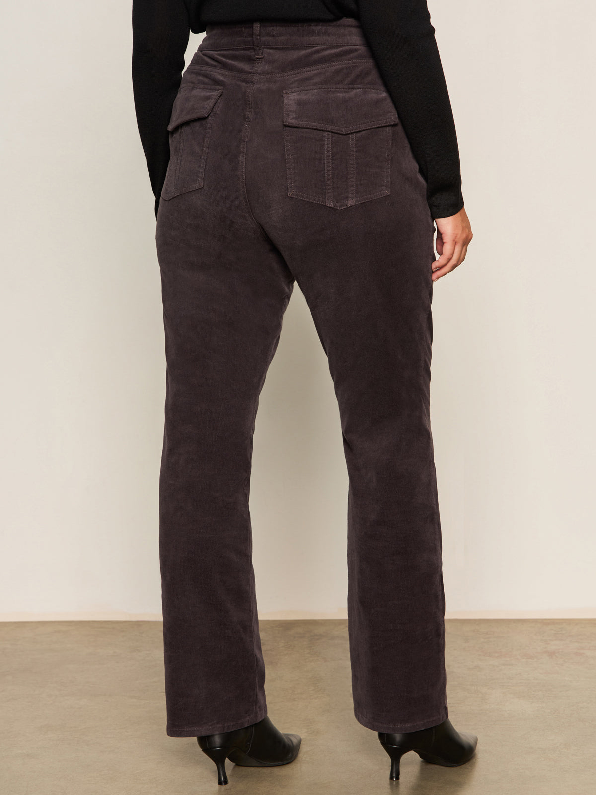 A person is shown from the back wearing Sanctuary Clothing's corduroy Hayden bootcut pant in semi sweet, featuring high-waisted style and patch back pockets, paired with black heeled boots and a black top against a plain background.