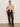 A woman stands against a plain wall wearing the Sanctuary Clothing fade into you ruffle blouse in fawn, paired with high-waisted black pants and black heeled ankle boots.