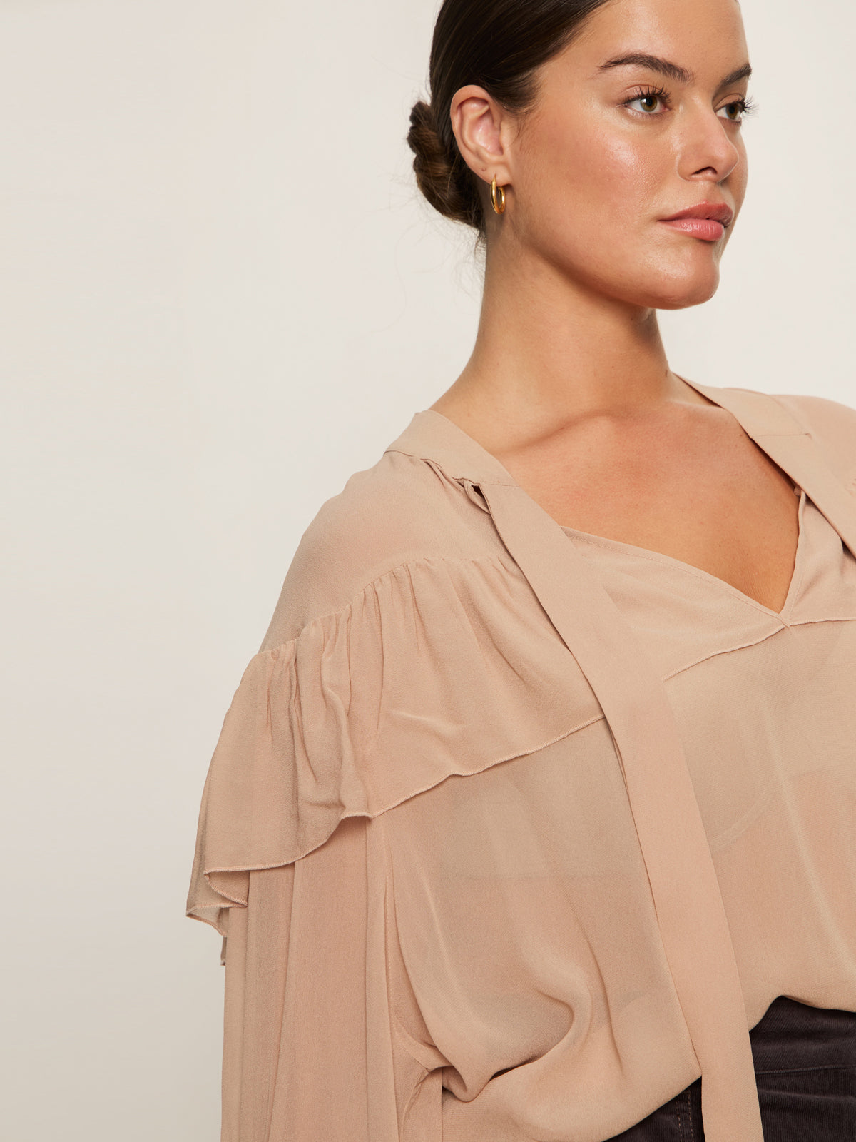 A woman with her hair in a low bun wears the Sanctuary Clothing "fade into you ruffle blouse" in fawn—a light beige, sheer blouse with ruffles, a V-neckline, and long, loose sleeves—while gazing to the side against a plain background.