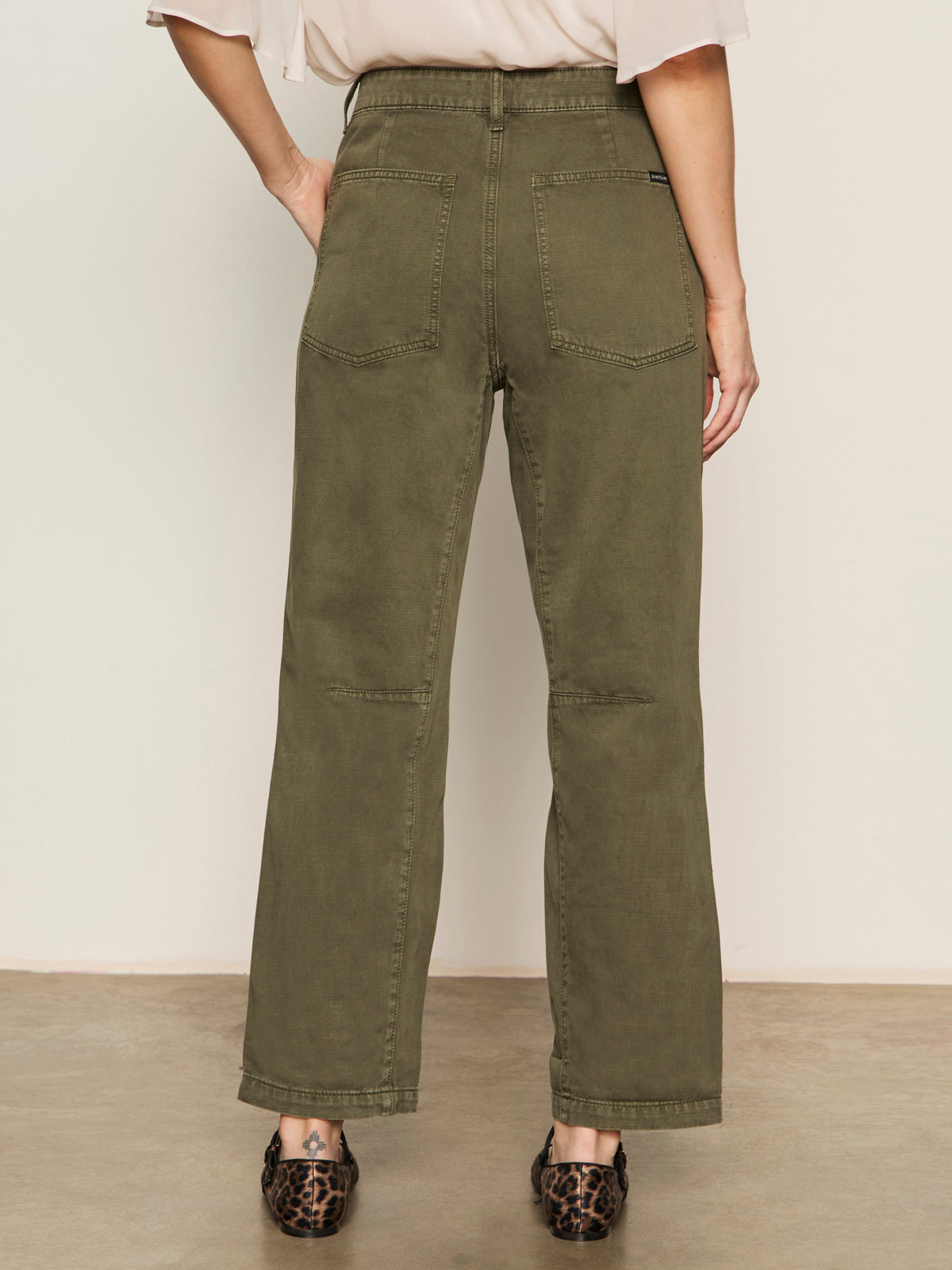 A person stands indoors, back facing camera, wearing Sanctuary Clothing™s boyfriend utility high rise pant in kalamata, paired with a light top and leopard print flats.