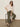 A woman in dark sunglasses, a loose white blouse, and Sanctuary Clothing™s "boyfriend utility high rise pant kalamata" sits on a black stool with one hand in her pocket, wearing leopard print flats. The background is plain and light-colored.