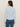 A woman with long brown hair is seen from the back wearing the Sanctuary Clothing dreamgirl lace button up top in blue powder, styled with blue jeans. The collared, textured top features a subtle geometric pattern against a beige background.