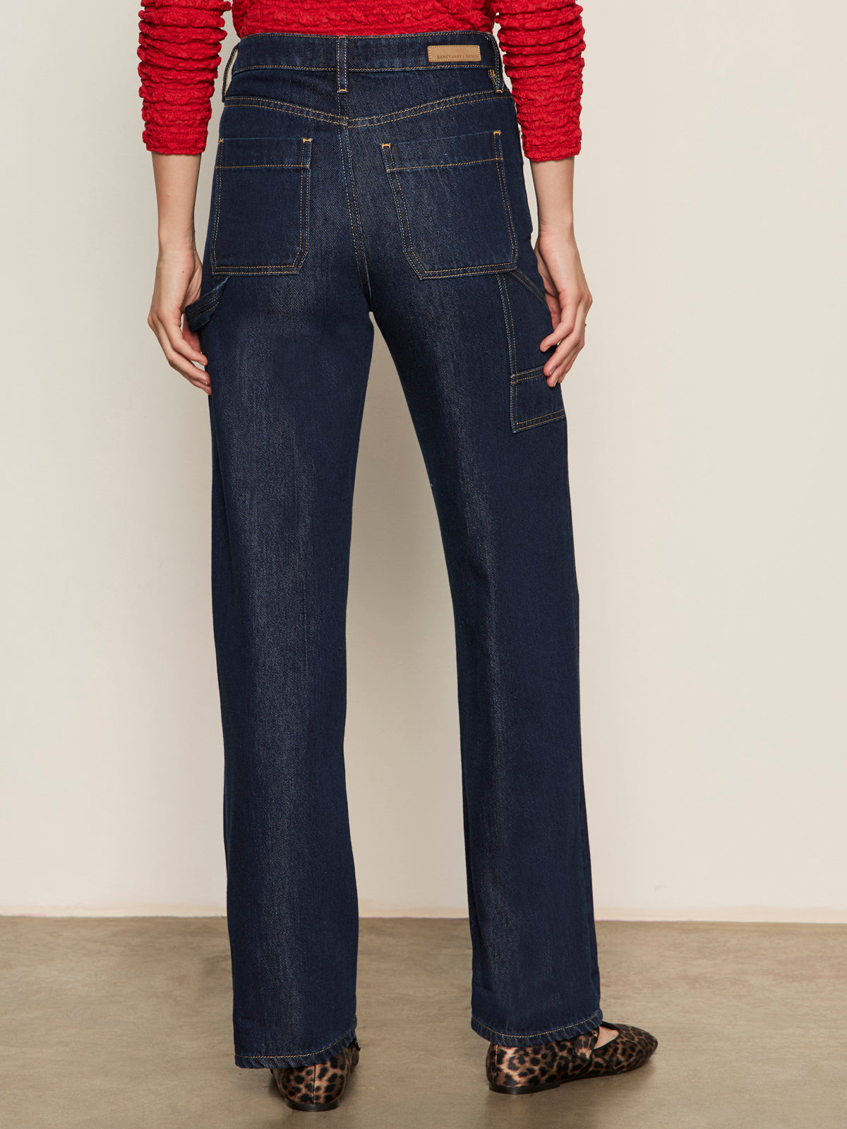 A person stands facing away, wearing Sanctuary Clothing soleil 90s straight jeans indigo dark raw rinse with large back pockets, a red long-sleeve top, and leopard-print flats.