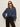 A woman with long brown hair wears black sunglasses, black pants, and the Sanctuary Clothing personal best blouse moonshadow�a loose-fitting blue denim button-up�standing against a plain light background with one hand in her pocket.