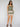 A woman with long brown hair and a neutral expression stands against a plain background wearing the Sanctuary Clothing spring breaker short mezzo sage romper with a square neckline, shorts, and brown sandals. Her hands are in her pockets.
