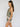 A woman stands in profile wearing the Sanctuary Clothing button back cami mezzo sage”a green sleeveless top with an ornate floral pattern and white geometric trim”against a plain light background.