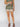 A person stands against a light background wearing Sanctuary Clothing's "Spring Breaker Short Mezzo Sage" set with green floral patterns and brown sandals. Their head is not visible in the image.