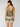 A woman with long brown hair faces away, wearing the Sanctuary Clothing button back cami mezzo sage, a sleeveless green top with a square neckline, white trim, and matching shorts featuring a pink and cream floral-like pattern.