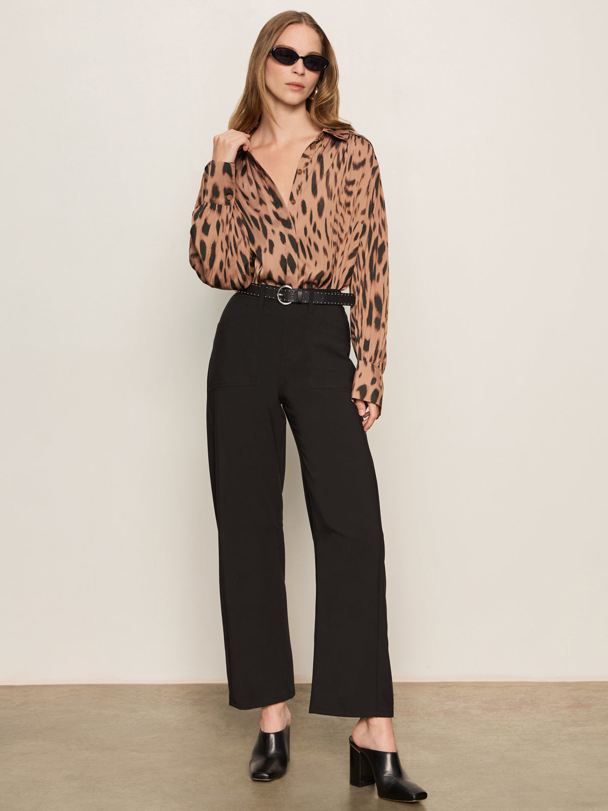 A woman stands against a plain, light wall wearing sunglasses, a brown animal print blouse, black heeled mules, and the Sanctuary Clothing charter gab barrel pant in black, with one hand touching her collar.