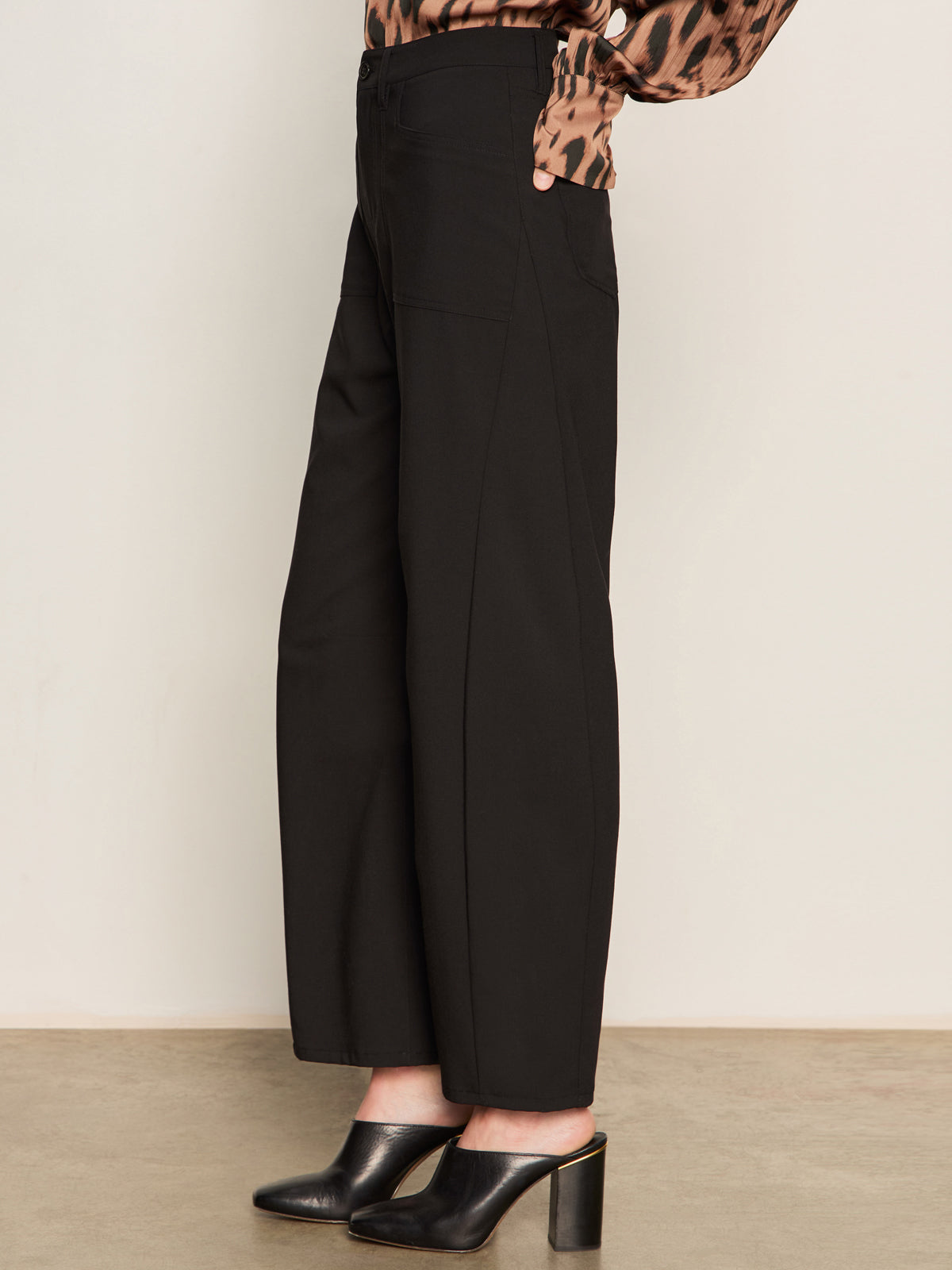 A person wearing the Sanctuary Clothing charter gab barrel pant in black stands on a light brown floor against a plain white wall. The side-view image, cropped from mid-torso down, shows the pants styled with a blouse and black heeled mules.