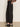 A person wearing the Sanctuary Clothing charter gab barrel pant in black stands on a light brown floor against a plain white wall. The side-view image, cropped from mid-torso down, shows the pants styled with a blouse and black heeled mules.