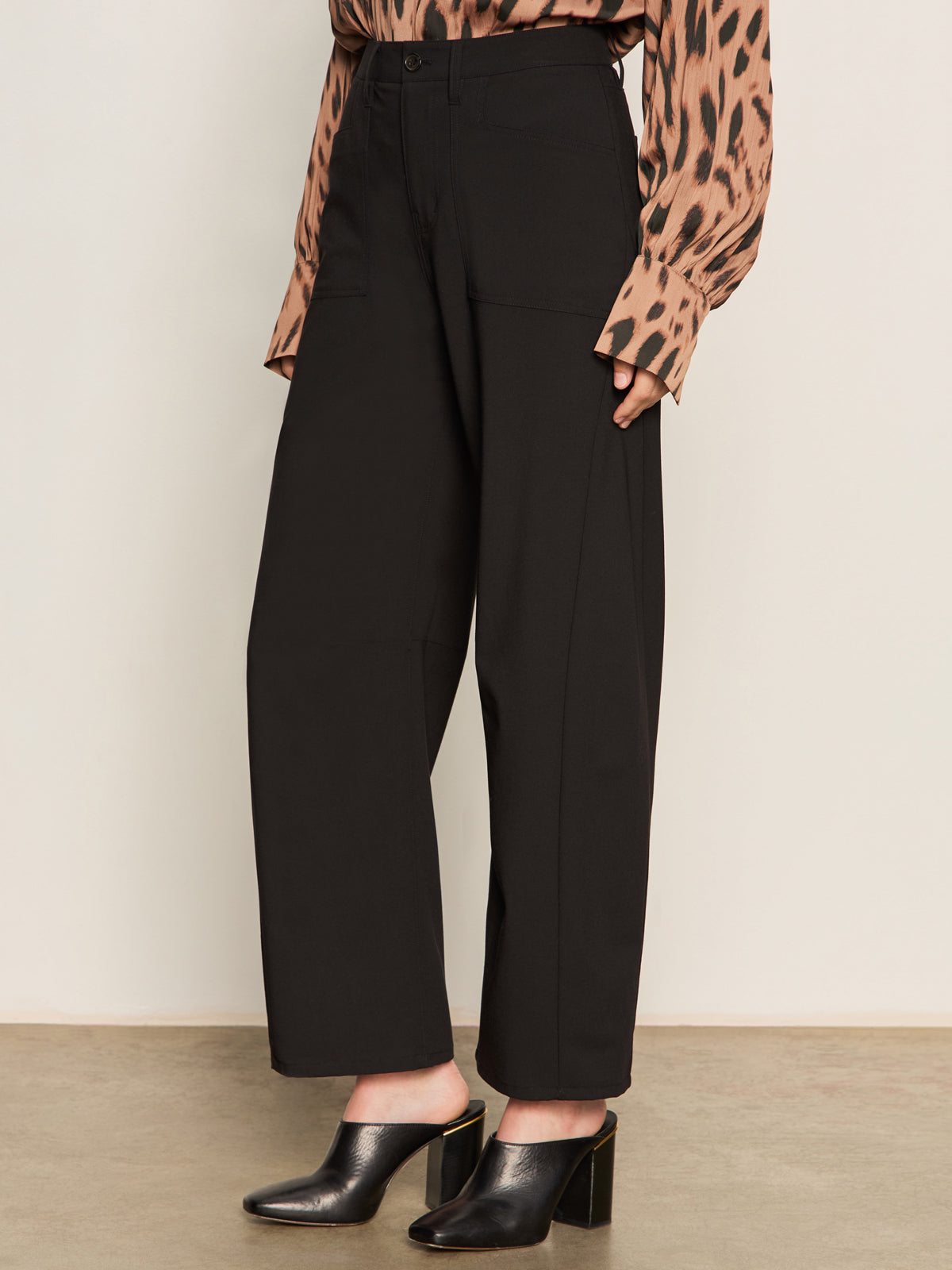 A person wearing Sanctuary Clothing's Charter Gab Barrel Pant in black, paired with a long-sleeved animal print blouse and black heeled mules, stands on a beige floor against a plain light-colored wall.
