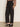 A person wearing Sanctuary Clothing's Charter Gab Barrel Pant in black, paired with a long-sleeved animal print blouse and black heeled mules, stands on a beige floor against a plain light-colored wall.
