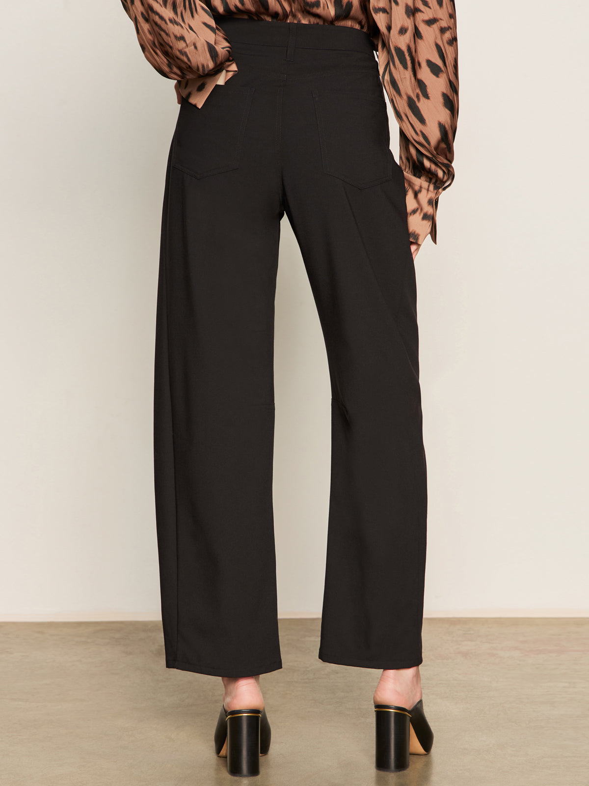 Indoors, a person faces away wearing Sanctuary Clothing's charter gab barrel pant in black, a brown and black patterned long-sleeve blouse, and black open-back block-heeled shoes.