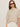 A woman with long brown hair wears black sunglasses, white pants, and the Sanctuary Clothing perfect sweater tee in heather pearl. She stands against a plain light background, looking slightly to the side with a neutral expression.