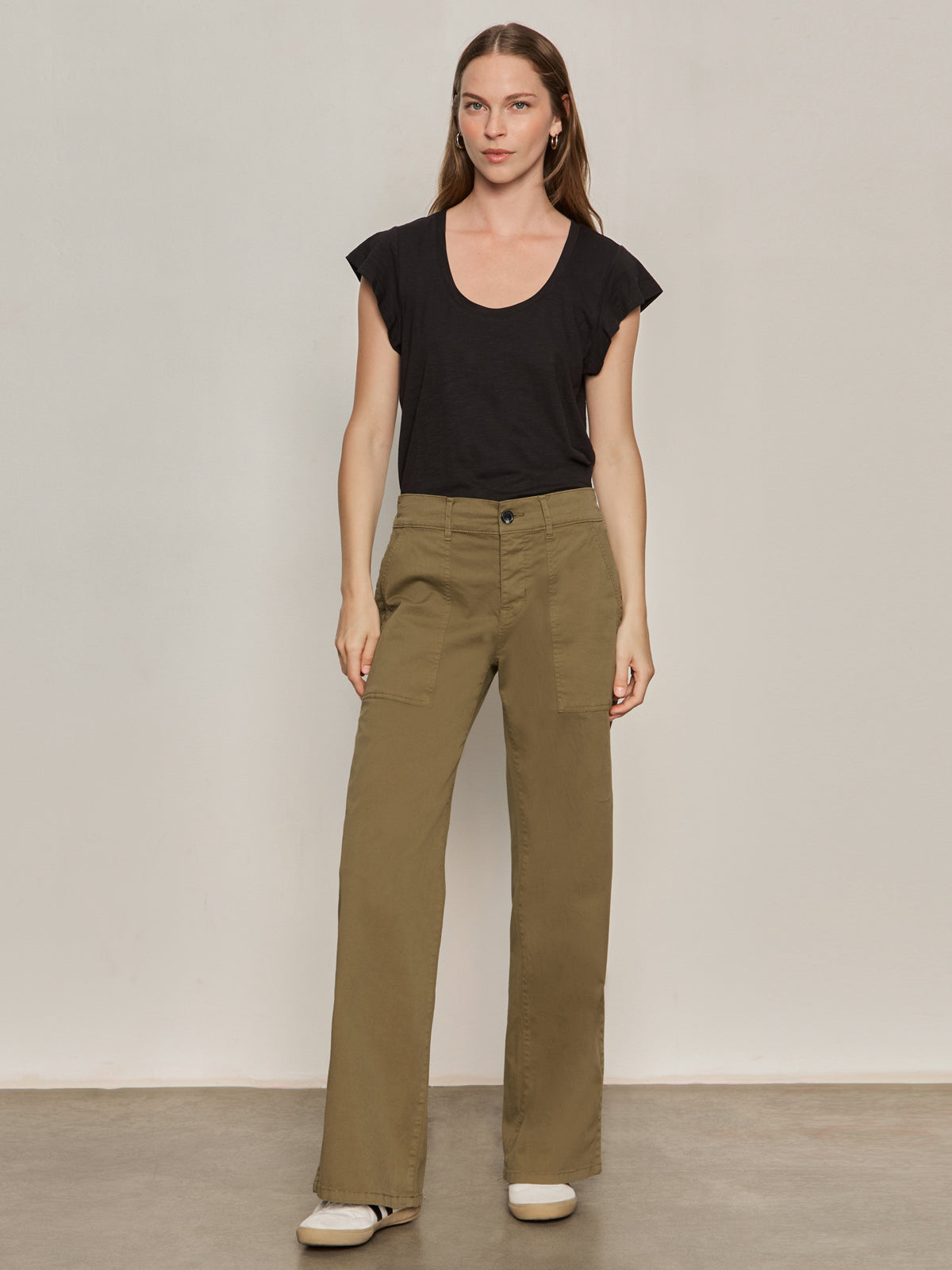 A woman stands against a plain light wall, wearing Sanctuary Clothing's cruiser full length pant in dirty martini, a black short-sleeve top, and white sneakers. Her hair is down and her expression is neutral.