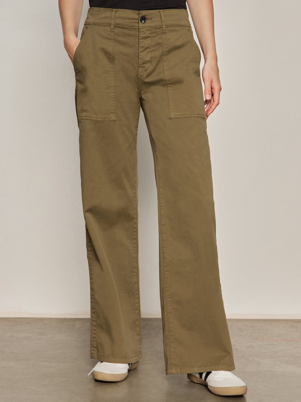 A person wearing Sanctuary Clothing’s cruiser full length pant in dirty martini, featuring wide legs and large front pockets, stands on a gray floor. Only the lower half and their white sneakers are visible.