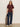 A woman stands against a plain wall wearing the Sanctuary Clothing shirred waist dolman tee in chicory, paired with dark blue wide-leg jeans and white sneakers. She holds a leopard print jacket and has straight, light brown hair.
