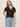 A woman with long, light brown hair wears Sanctuary Clothing’s notch neck tee in black, tucked into light blue jeans with a black belt. She stands against a plain, light-colored background, looking slightly past the camera.