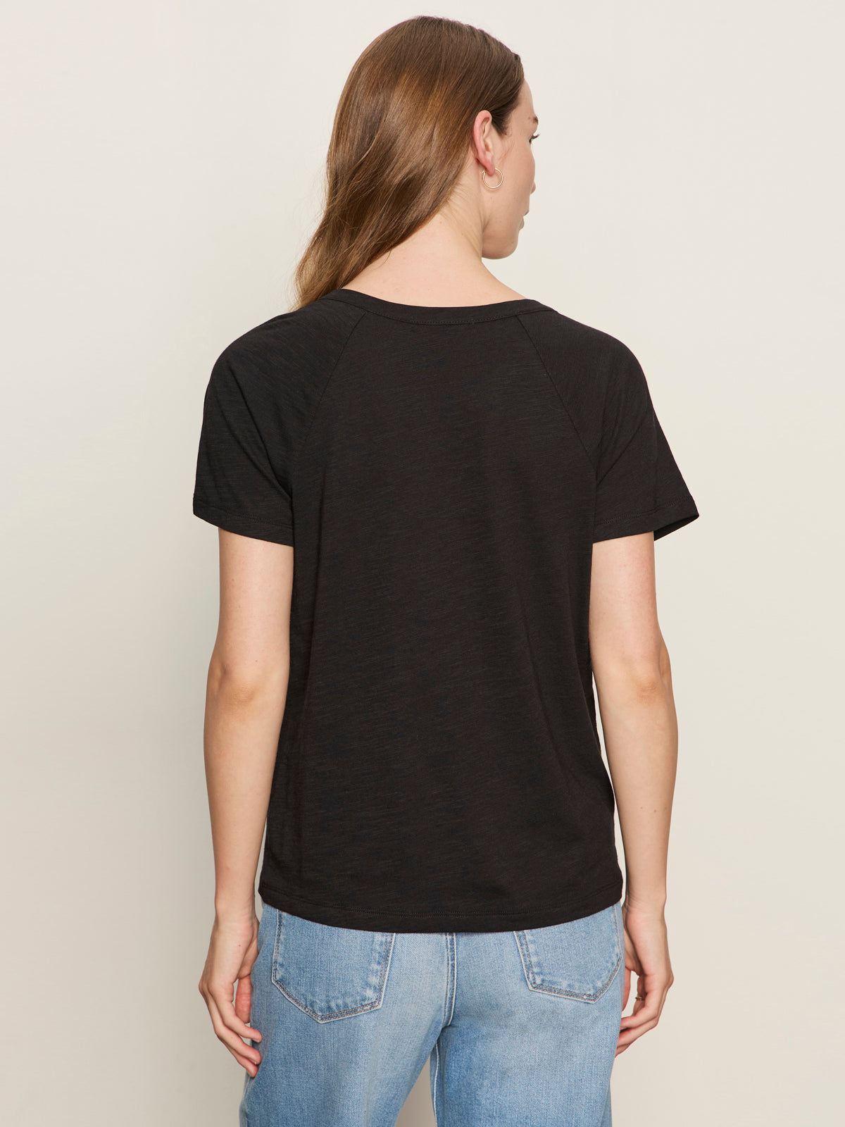 A person with long brown hair is seen from the back against a light background, wearing the Sanctuary Clothing notch neck tee in black and light blue jeans.