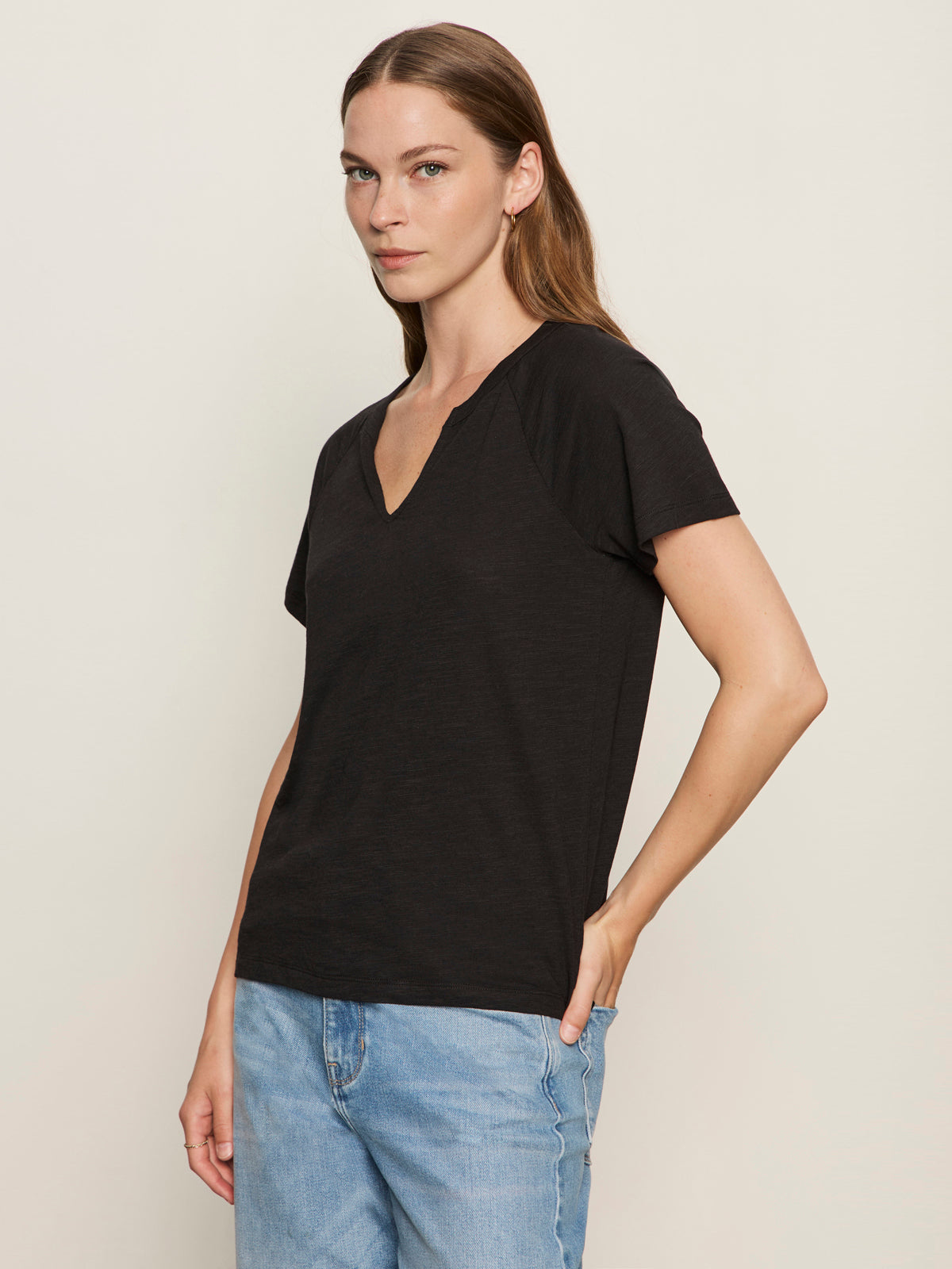 A person with long brown hair wears the Sanctuary Clothing notch neck tee in black and light blue jeans, standing against a plain light background with one hand in their pocket.