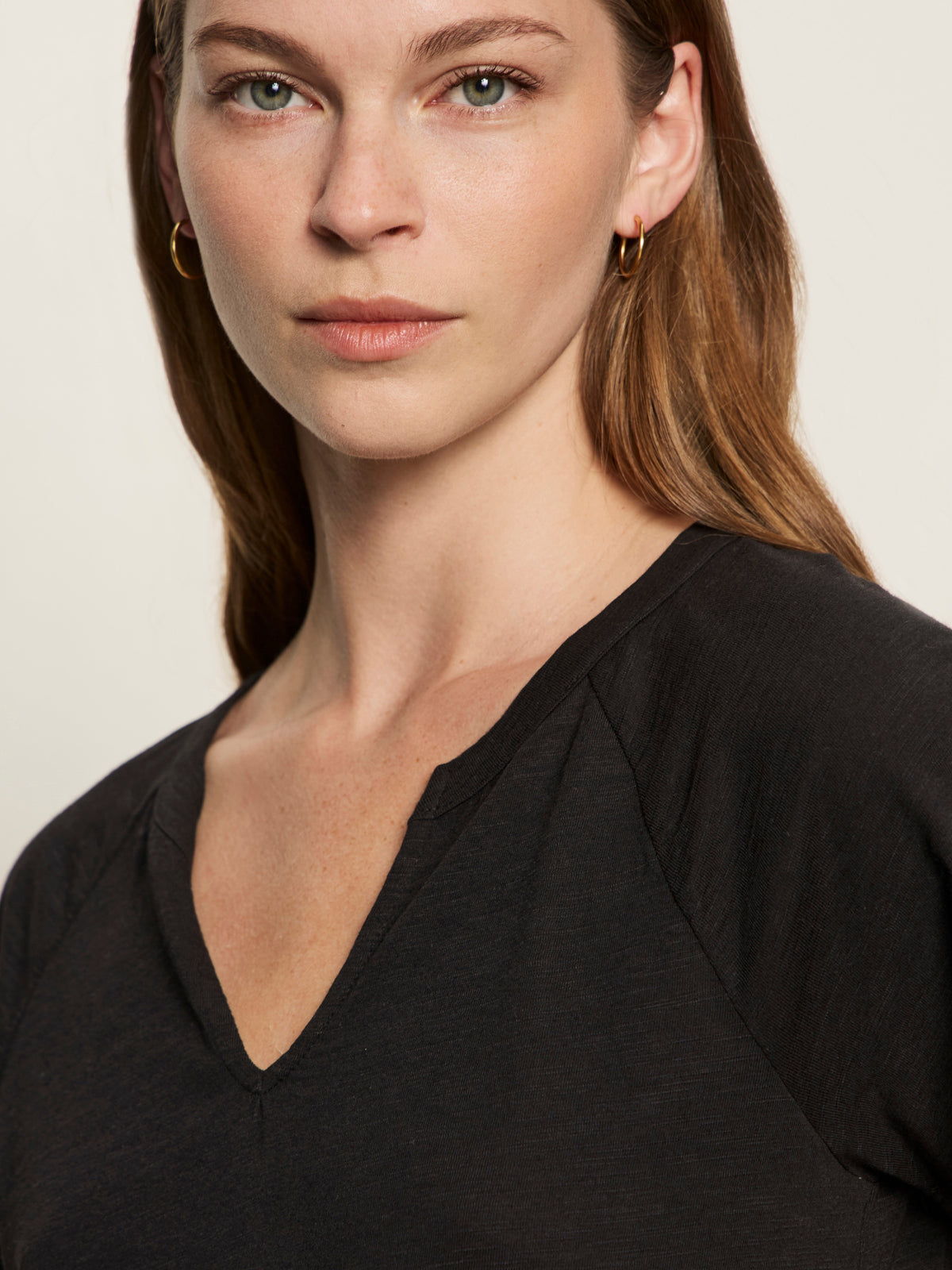 A woman with light skin, straight brown hair, and green eyes wears small gold hoops and a black notch neck tee by Sanctuary Clothing, looking confidently at the camera against a neutral background.