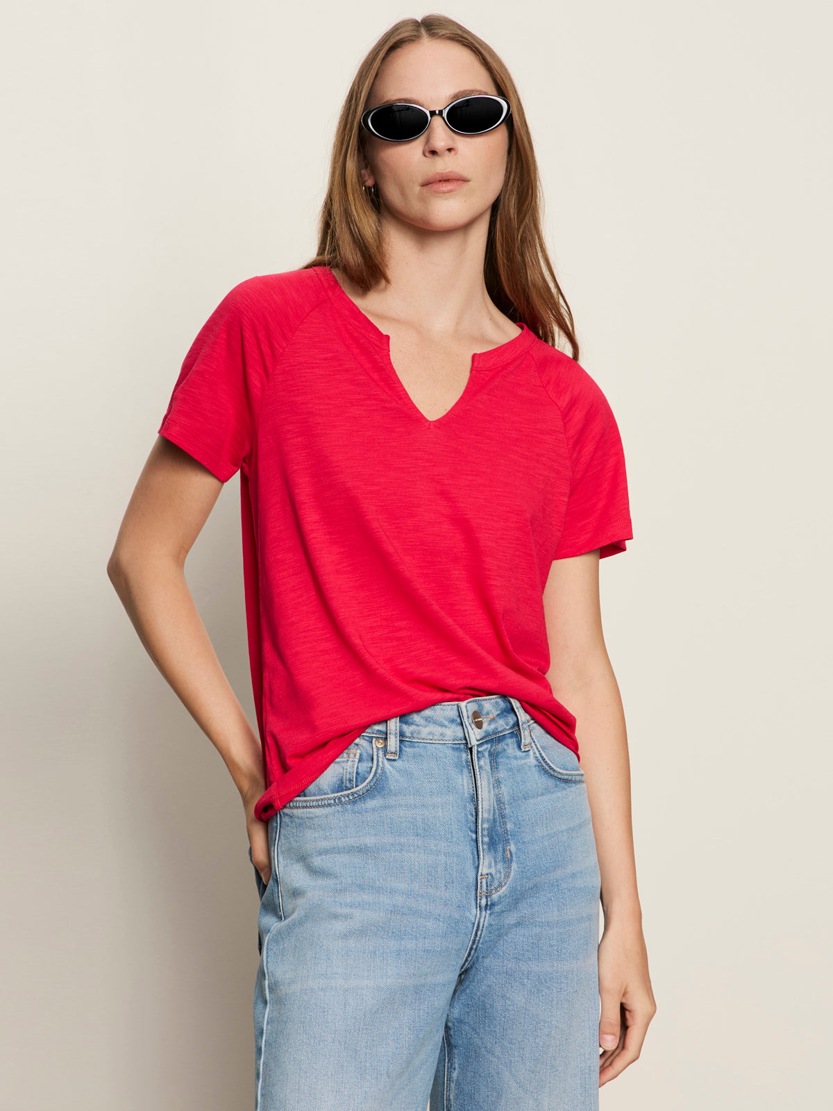 A woman with long brown hair wears the Sanctuary Clothing notch neck tee radiant, light blue jeans, and black oval sunglasses, standing with one hand in her pocket against a neutral background.