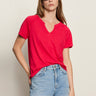 A woman with long brown hair wears the Sanctuary Clothing notch neck tee radiant, light blue jeans, and black oval sunglasses, standing with one hand in her pocket against a neutral background.