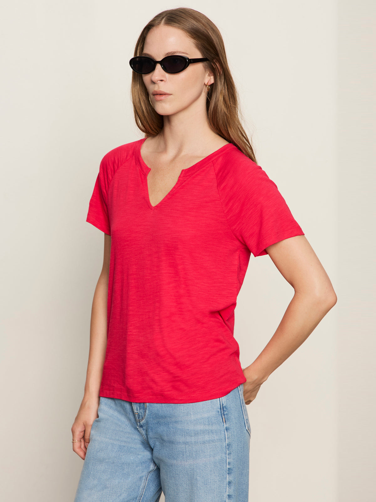 A woman wearing black sunglasses, light blue jeans, and a Sanctuary Clothing notch neck tee radiant stands against a plain beige background with one hand on her hip.
