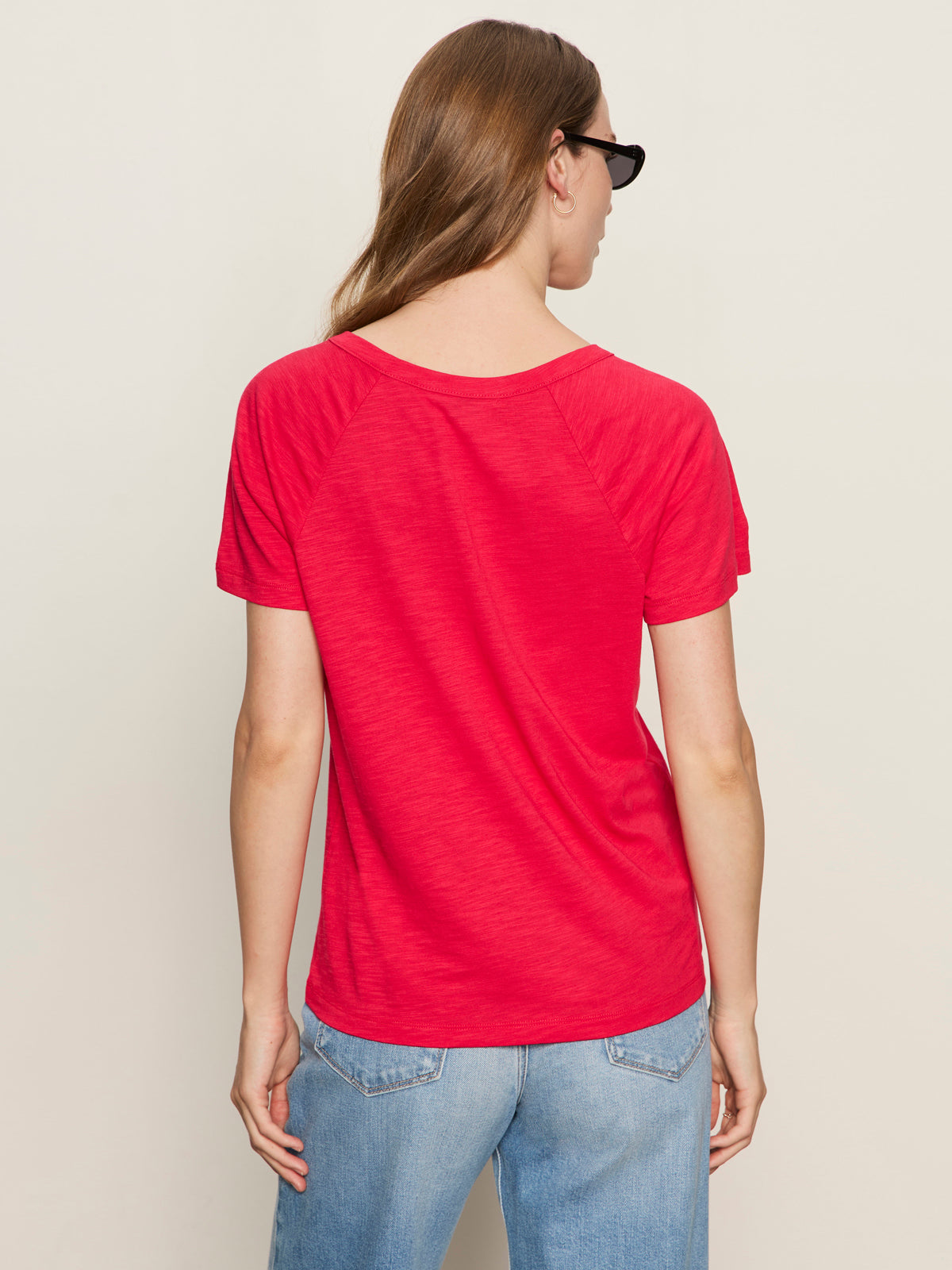A woman with long brown hair, wearing sunglasses and the Sanctuary Clothing notch neck tee radiant in bright red with light blue jeans, stands with her back to the camera against a plain light background.