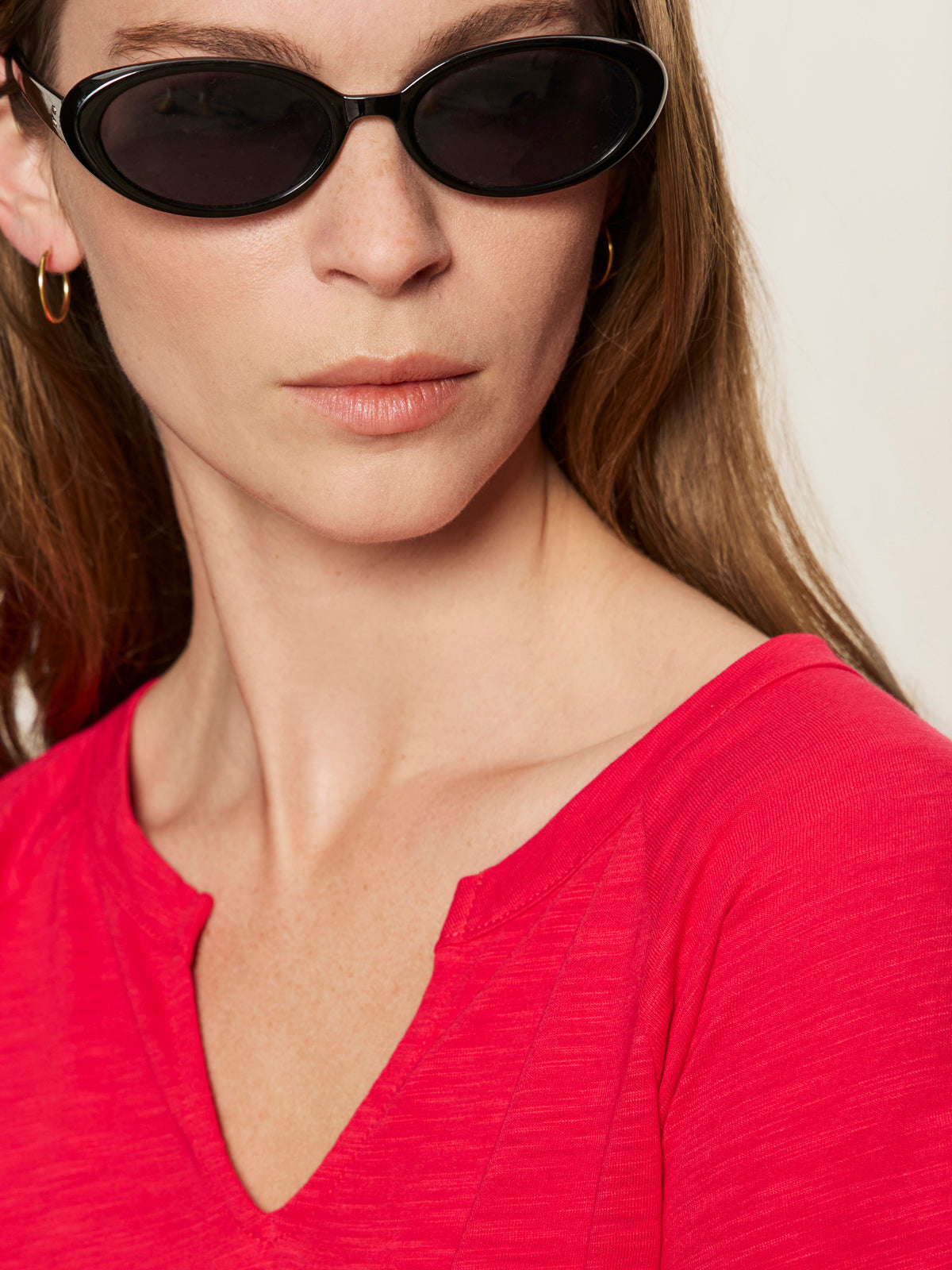 A woman with light skin and long brown hair wears black oval sunglasses, small gold hoop earrings, and the Sanctuary Clothing notch neck tee radiant in red. She looks slightly to the side against a neutral background.