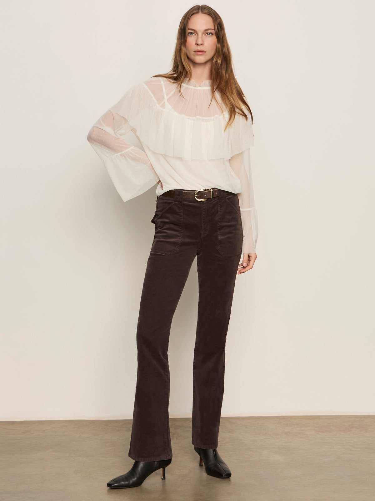 A woman stands against a plain wall wearing the ethereal blouse chalk by Sanctuary Clothing—a sheer white ruffled top—paired with high-waisted dark brown corduroy pants, black heeled boots, and a belt. Her long hair is down, with one hand at her side.