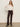 A woman stands against a plain wall wearing the ethereal blouse chalk by Sanctuary Clothing—a sheer white ruffled top—paired with high-waisted dark brown corduroy pants, black heeled boots, and a belt. Her long hair is down, with one hand at her side.