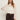 A woman with long brown hair wears Sanctuary Clothing’s “ethereal blouse chalk”—a sheer white ruffled top with flared sleeves—paired with dark high-waisted pants and a belt, standing against a plain light background.
