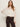 A woman with long brown hair wears Sanctuary Clothing’s “ethereal blouse chalk”—a sheer white ruffled top with flared sleeves—paired with dark high-waisted pants and a belt, standing against a plain light background.