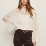 A woman with long brown hair wears Sanctuary Clothing’s “ethereal blouse chalk”—a sheer white ruffled top with flared sleeves—paired with dark high-waisted pants and a belt, standing against a plain light background.