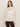 A woman with long brown hair models Sanctuary Clothing's "the ethereal blouse chalk," a sheer white ruffled, long-sleeve high-neck top, paired with dark pants, against a plain light background.