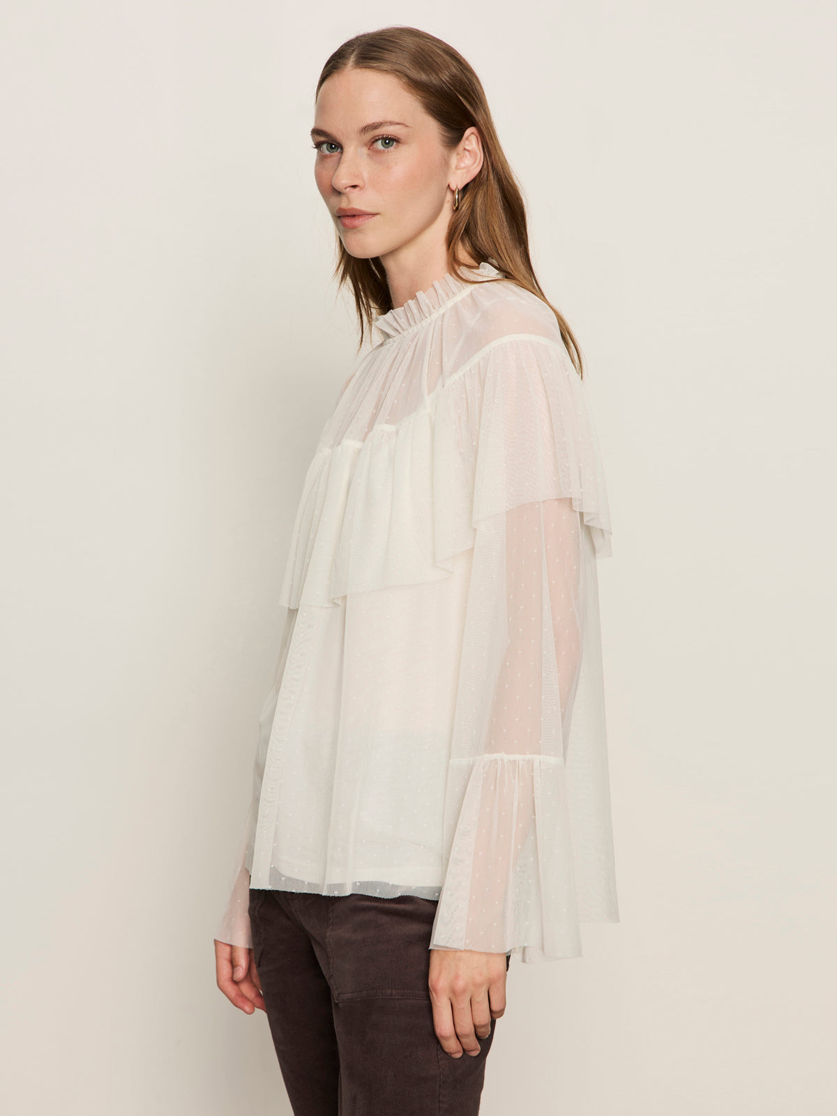A woman with long brown hair models Sanctuary Clothing's "the ethereal blouse chalk," a sheer white top with ruffled layers and bell sleeves, paired with dark pants, standing sideways against a plain light background.