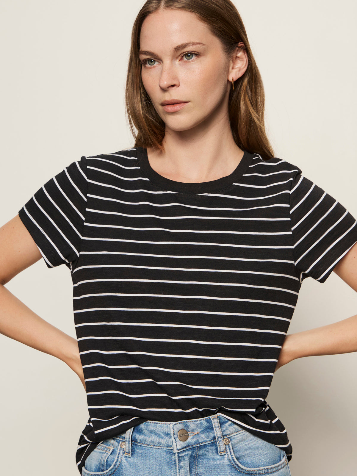 A woman with straight brown hair models the Sanctuary Clothing "the perfect tee white black stripe 5," a black T-shirt with white stripes, paired with light blue jeans, standing hands on hips against a plain light background.